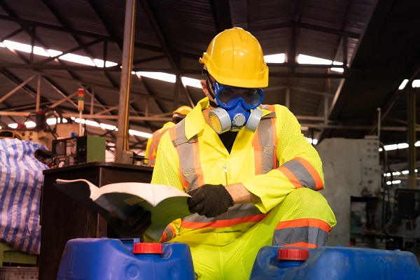 worker wearing protective mask, goggles, hard hat looks at containers of hazardous chemicals inside a factory.